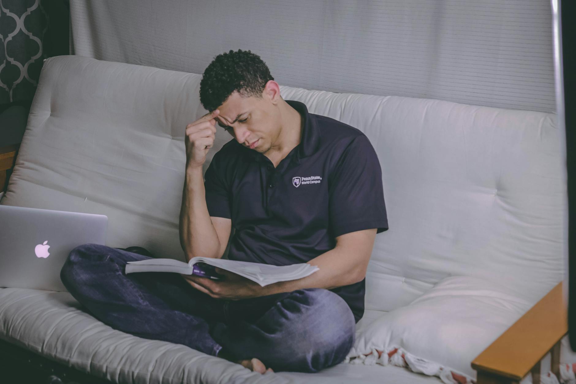 A student relaxing on a sofa, studying with a book and using a laptop indoors.